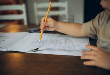 a childs hand draws at a wooden table with watercolors with a brush in an album
