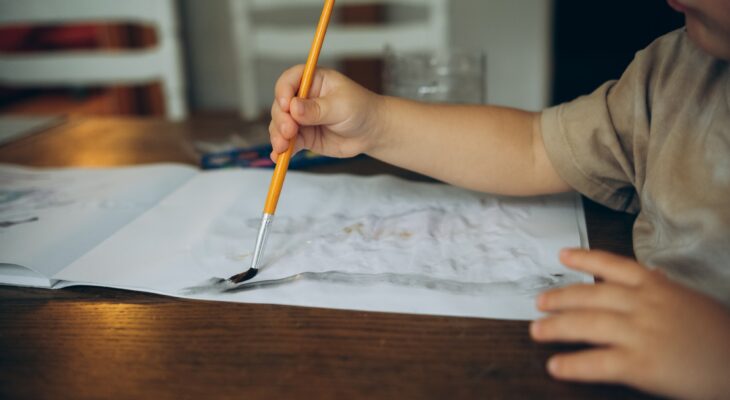 a childs hand draws at a wooden table with watercolors with a brush in an album