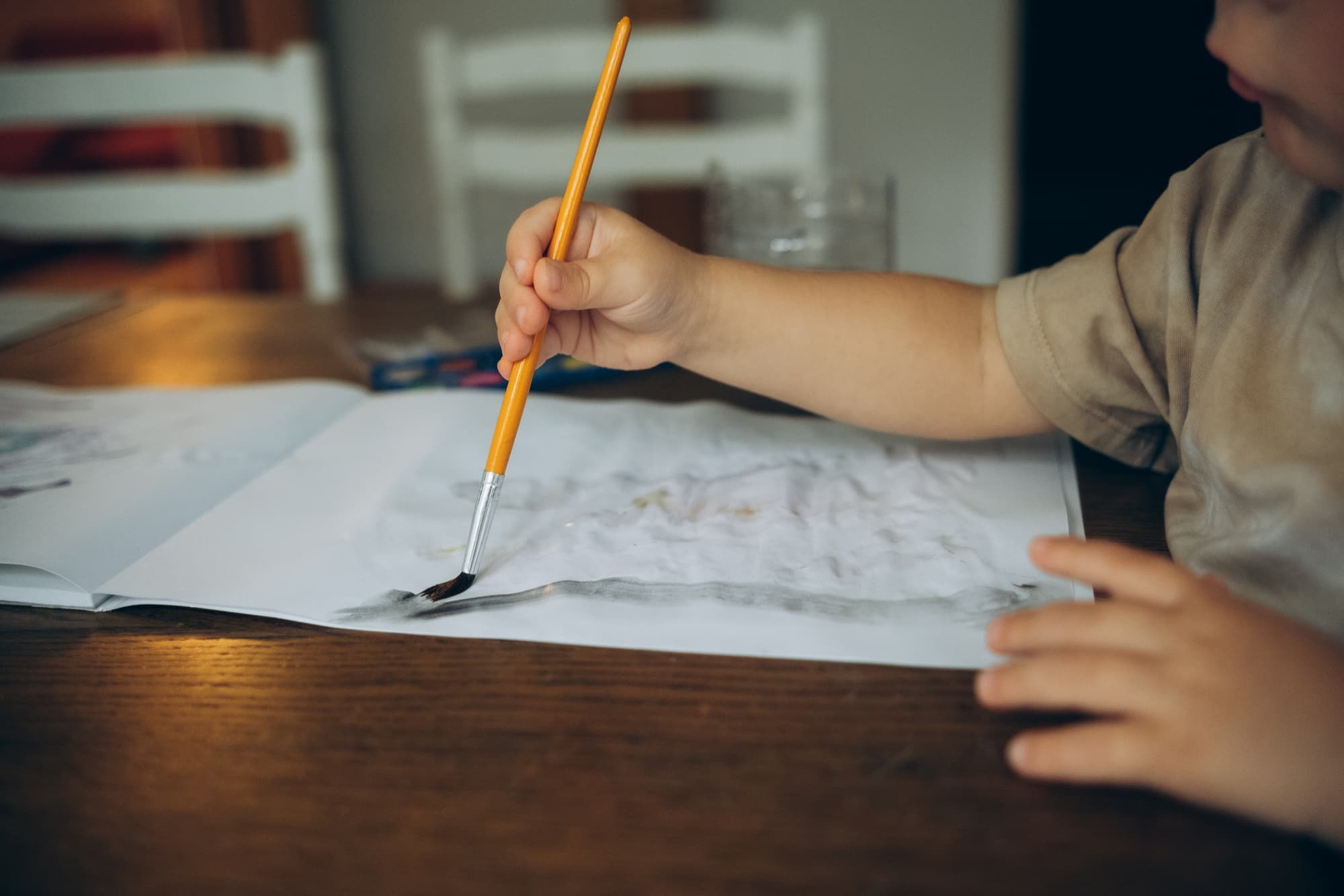 a childs hand draws at a wooden table with watercolors with a brush in an album