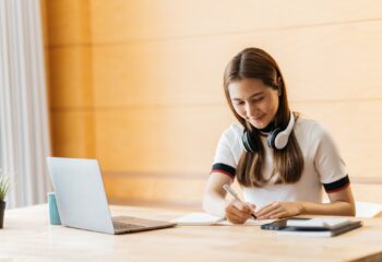 Happy asian businesswoman wearing headset make notes talk by conference call doing video chat on lap