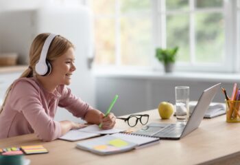 Laughing cute blonde european young girl in headphones study at home look at pc in kitchen interior