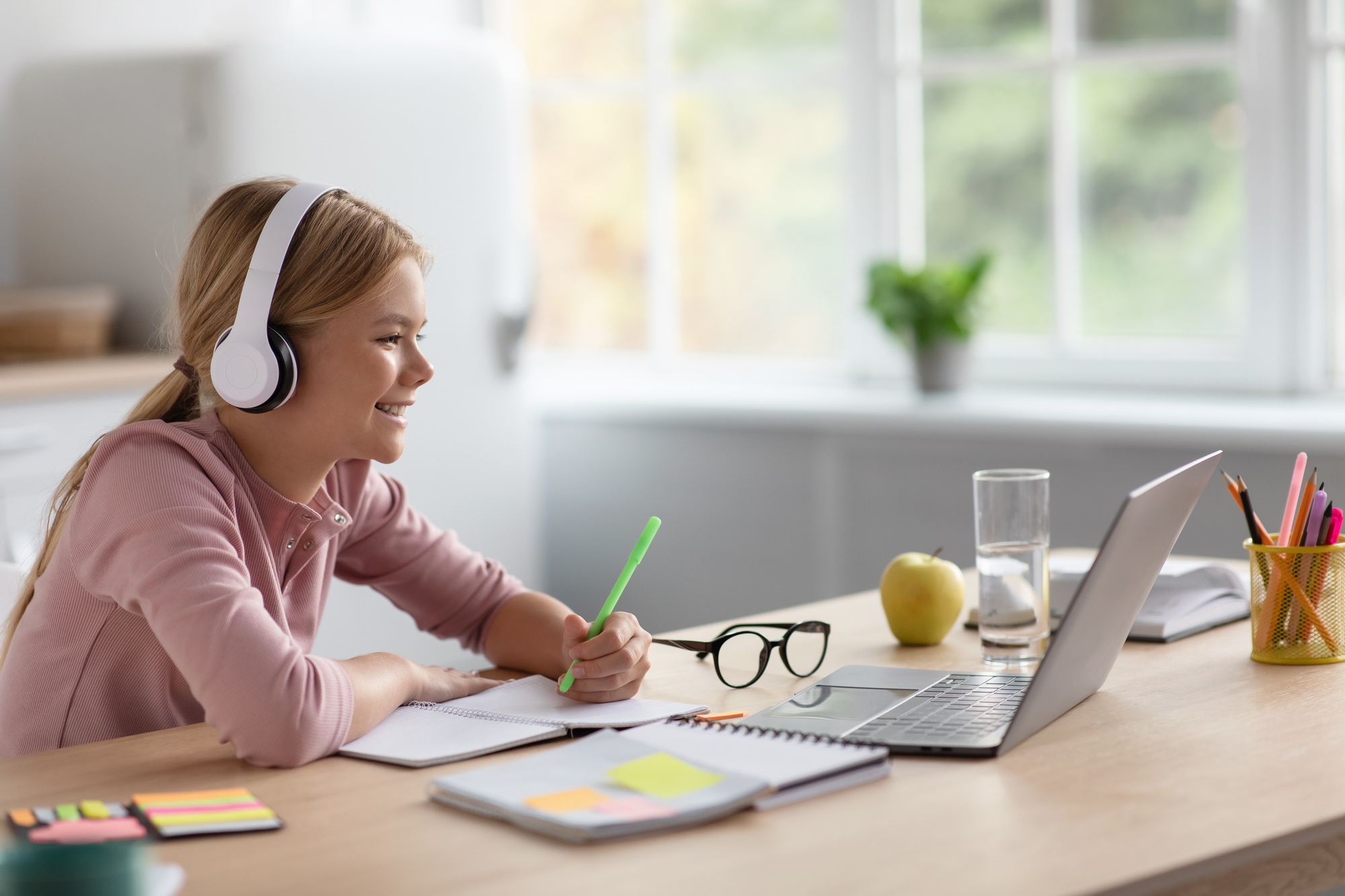 Laughing cute blonde european young girl in headphones study at home look at pc in kitchen interior